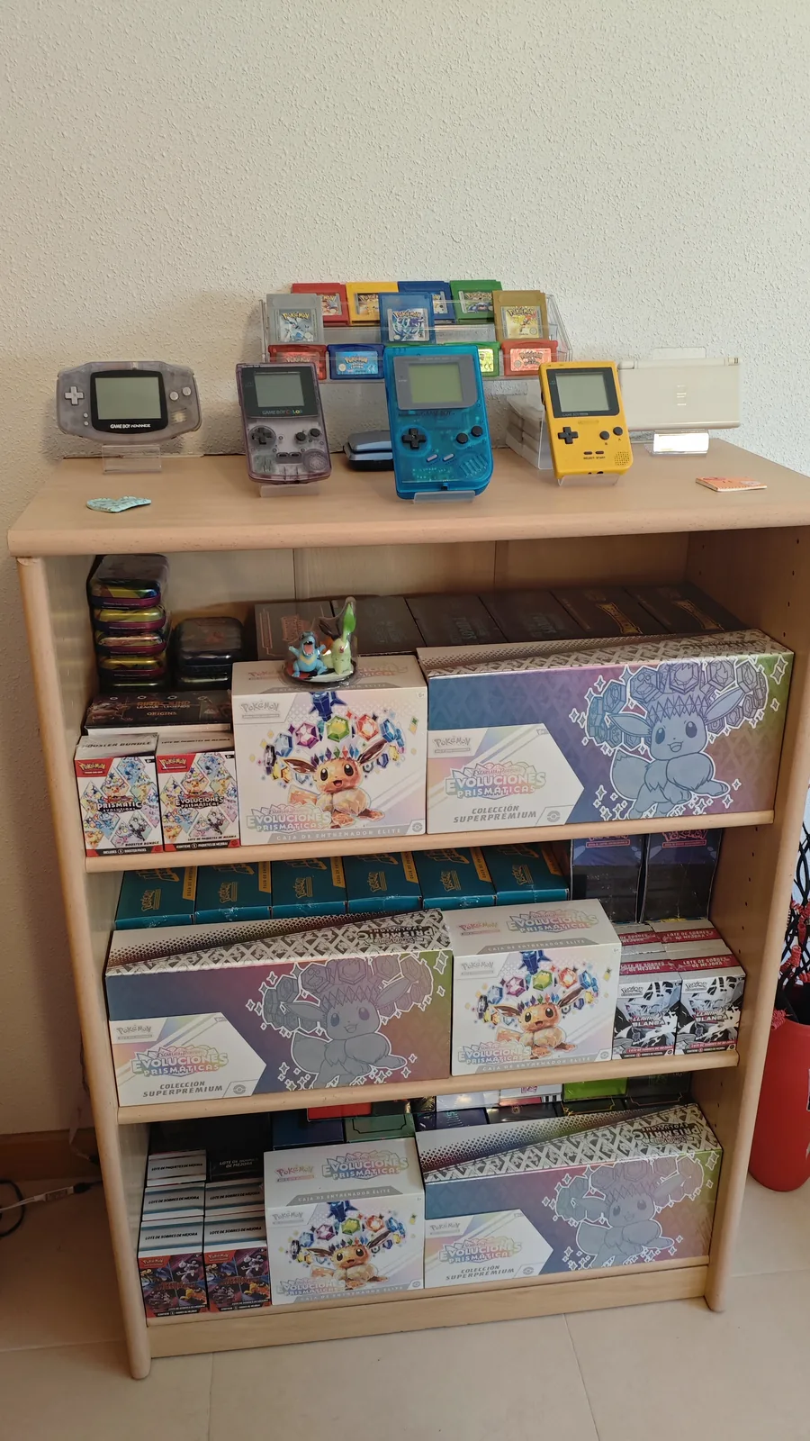 Estantería con colección de Pokémon TCG sellado (Evoluciones Prismáticas ETBs, Colección Superpremium, boosters) y consolas retro (Game Boy, GB Color, GB Pocket, GBA, DS Lite) con cartuchos originales